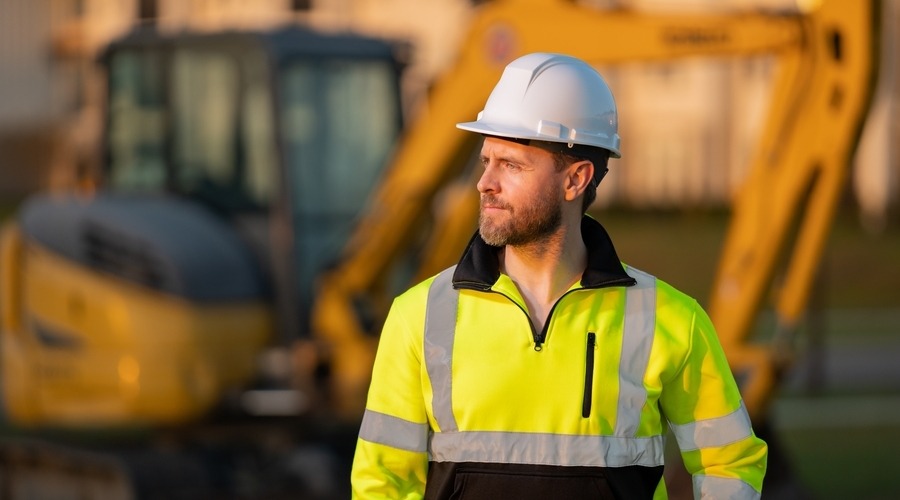 A construction site worker stands in front of an excavator.